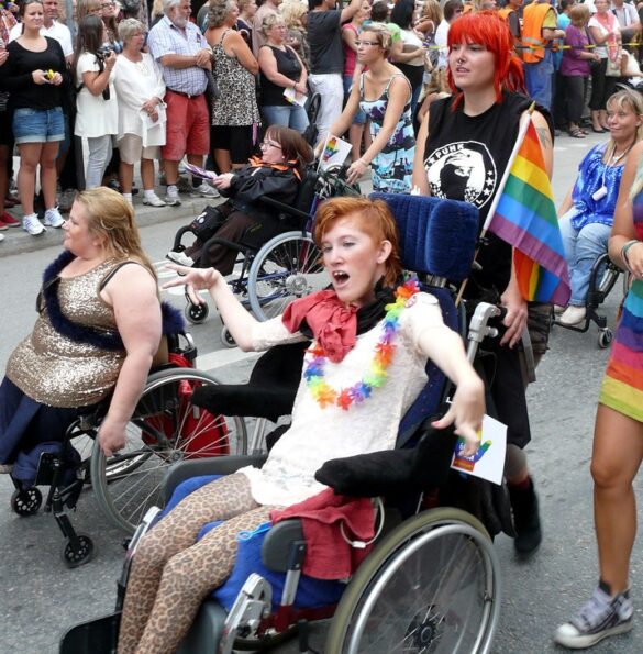 Alcune persone con disabilità sfilano in carrozzina durante una manifestazione, accompagnata da altre che le spingono. In primo piano una ragazza con decori arcobaleno sembra urlare. Sui lati della strada una folla le guarda incuriosite.